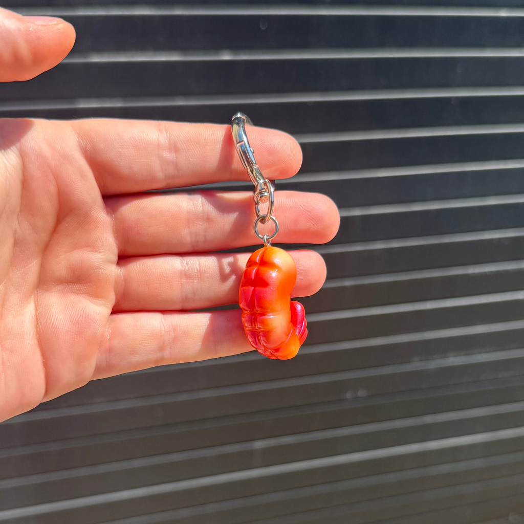 A hand holding a small orange and red shrimp -shaped charm with a silver lobster clasp, set against a dark gray slatted background.