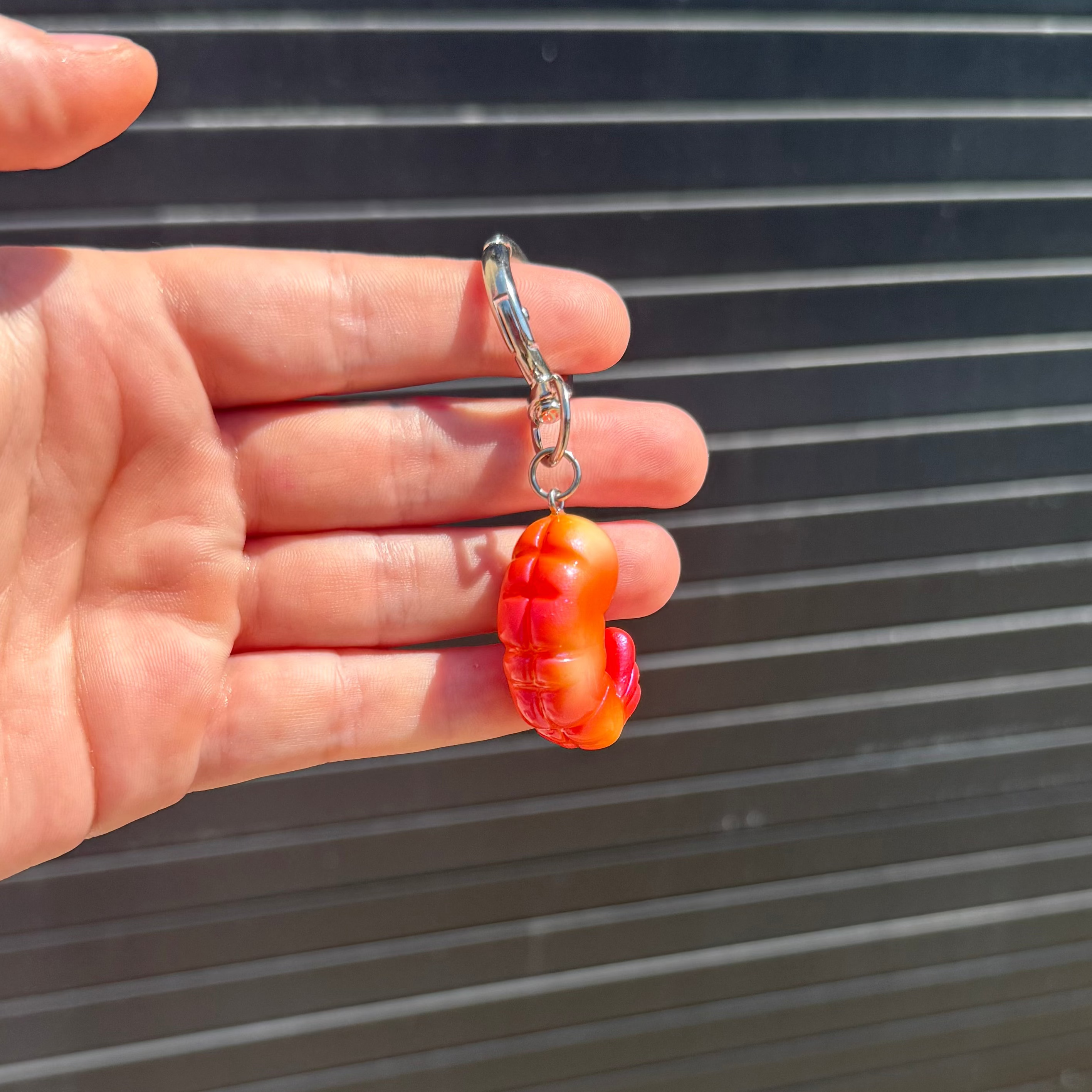 A hand holding a small orange and red shrimp -shaped charm with a silver lobster clasp, set against a dark gray slatted background.