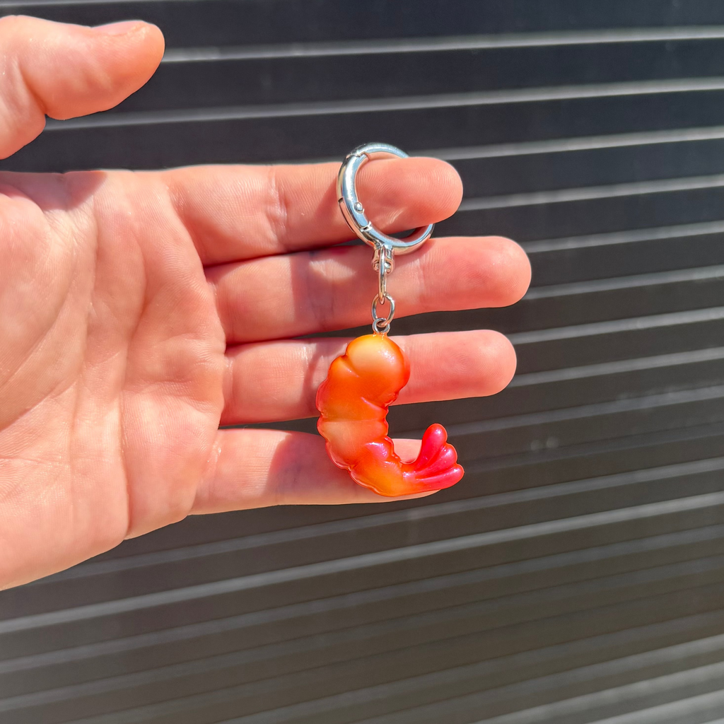 A hand holding a small keychain with a silver metal ring and a translucent orange and red shrimp-shaped charm against a dark corrugated metal background.