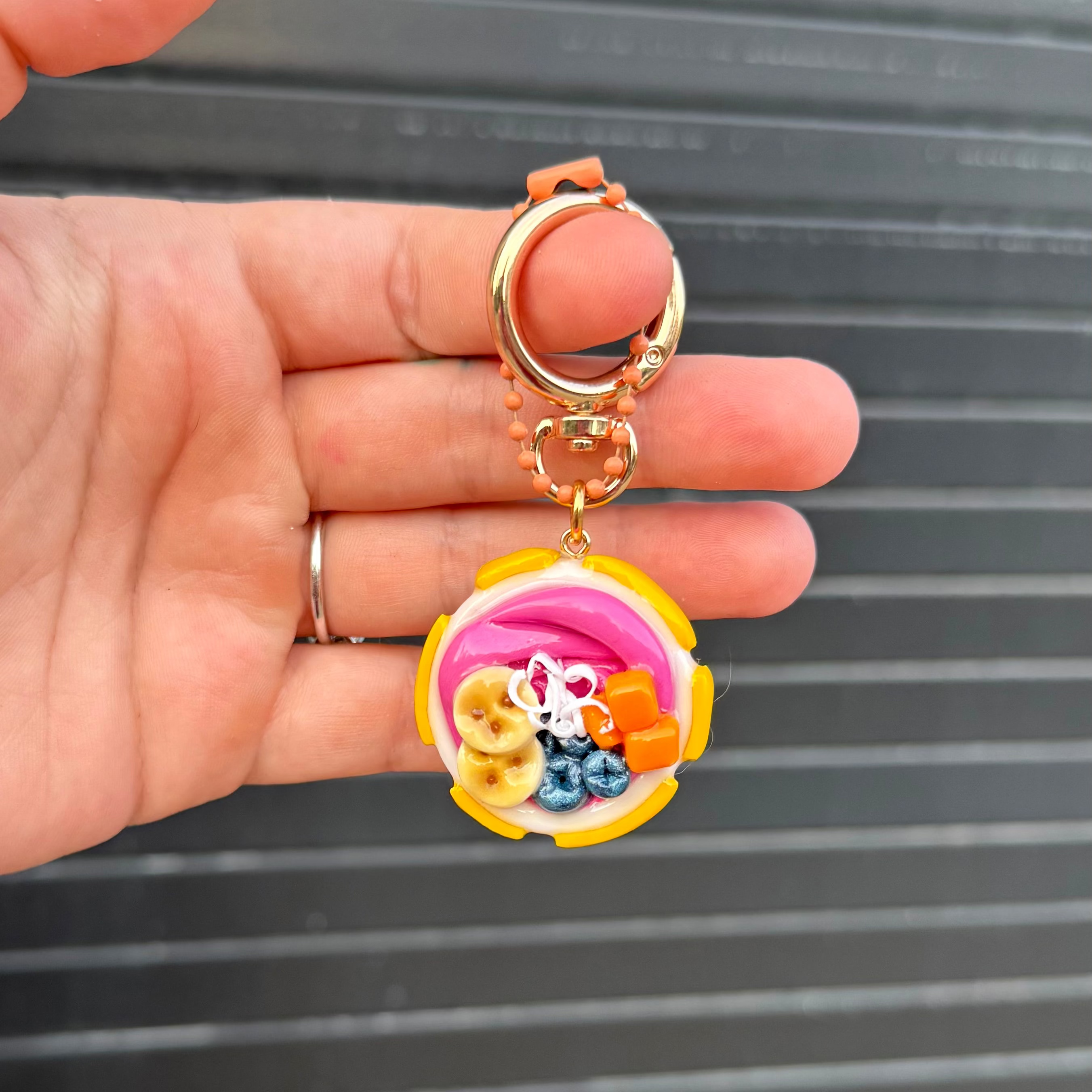A hand holding a small keychain with a gold-tone split ring and charm. The charm is a colorful, 3D sculpted piece resembling a pink and yellow bowl or cup filled with tiny decorative beads and charms, set against a dark gray slatted background.