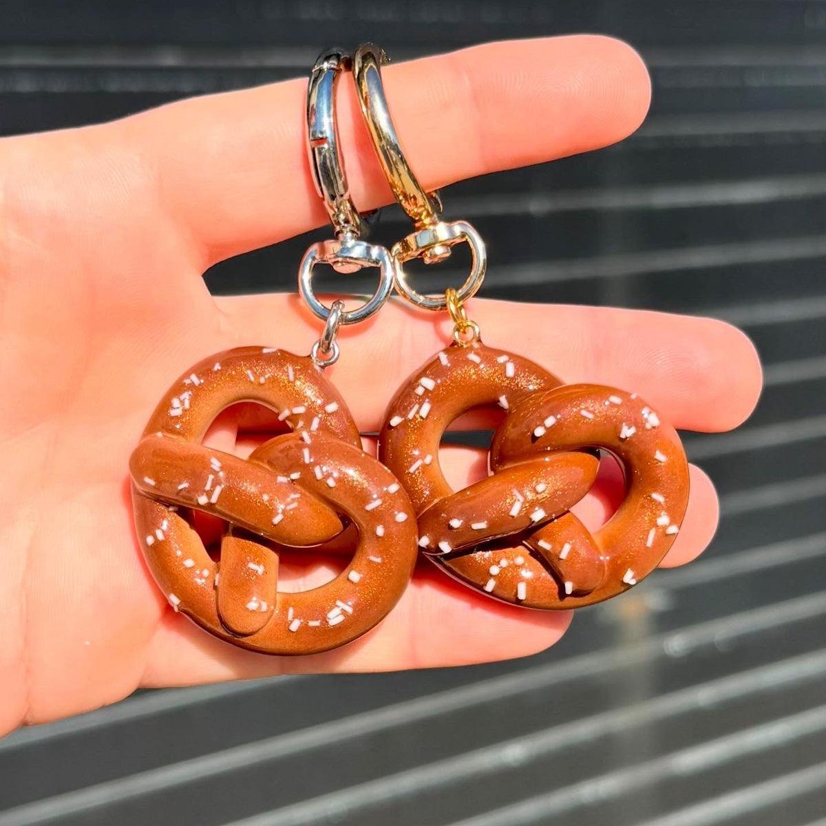 A pair of pretzel-shaped keychains held in a person's hand. The earrings have a glossy brown pretzel body with white salt details and silver-toned fishhook ear wires.