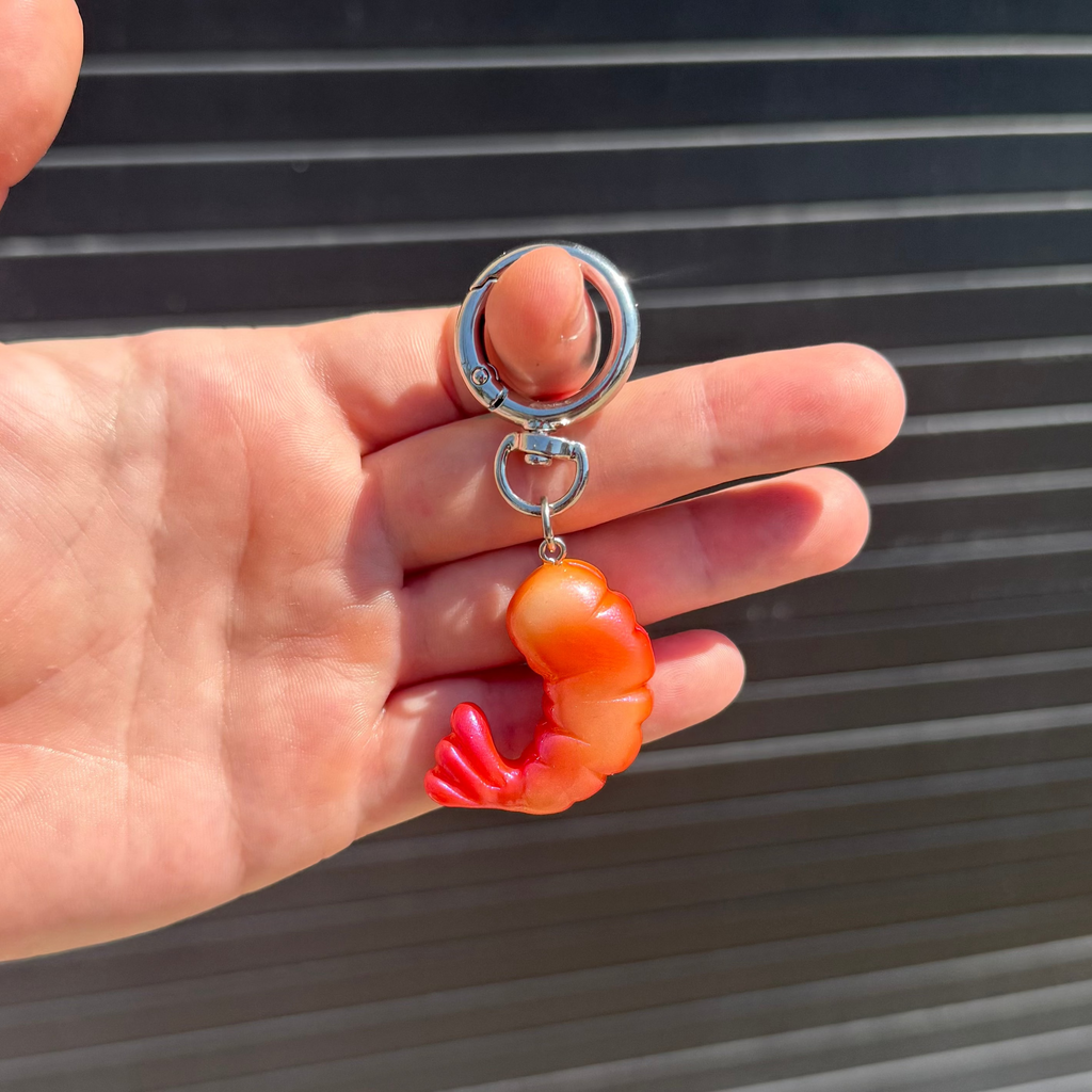 A hand holding a silver split keyring with a small shrimp-shaped charm that transitions from orange to red, set against a dark horizontal slatted background.