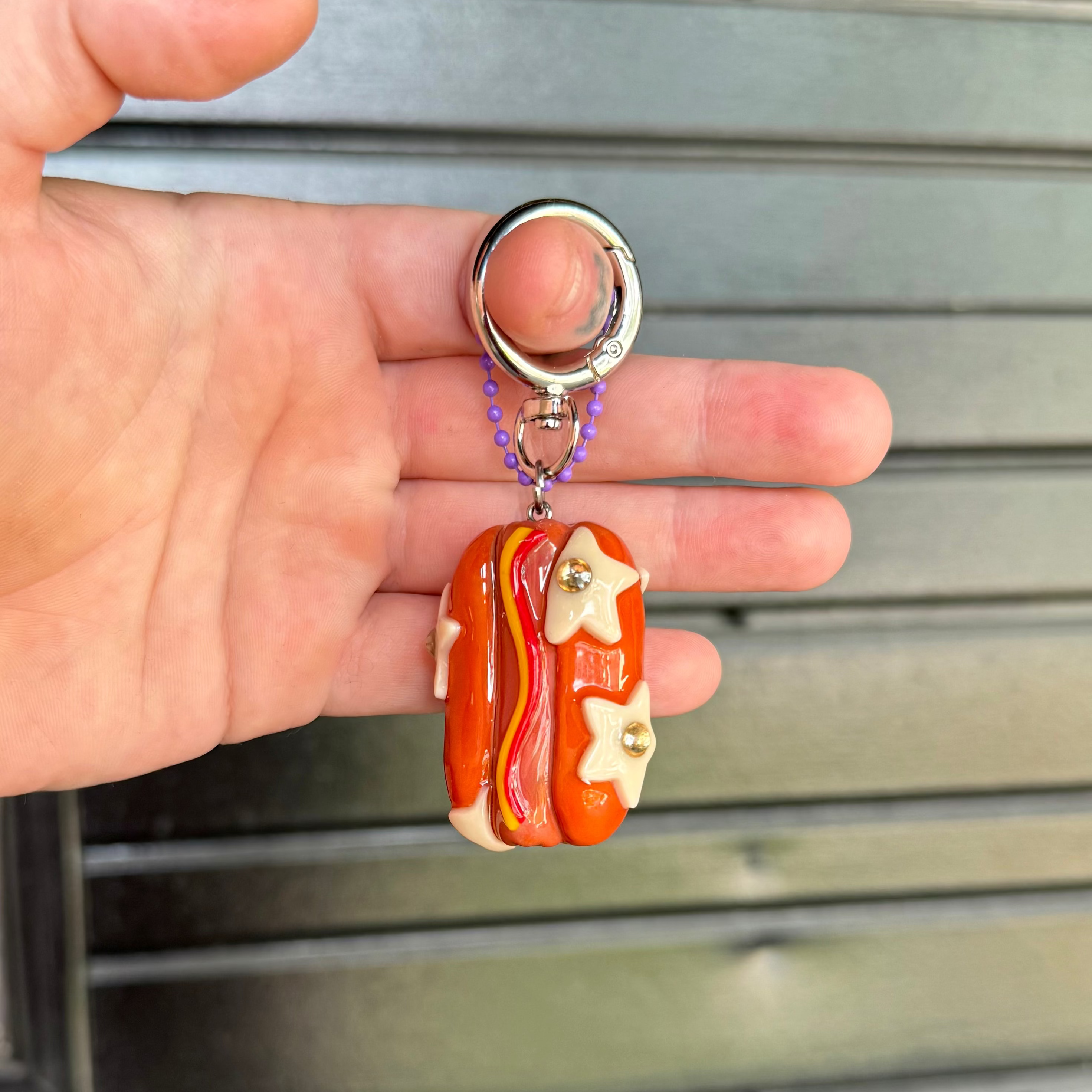 A hand holding a small keychain shaped like a hot dog, featuring a glossy orange-red bun with yellow stars, ketchup and mustard, and a silver key ring against a gray metal shutter background.