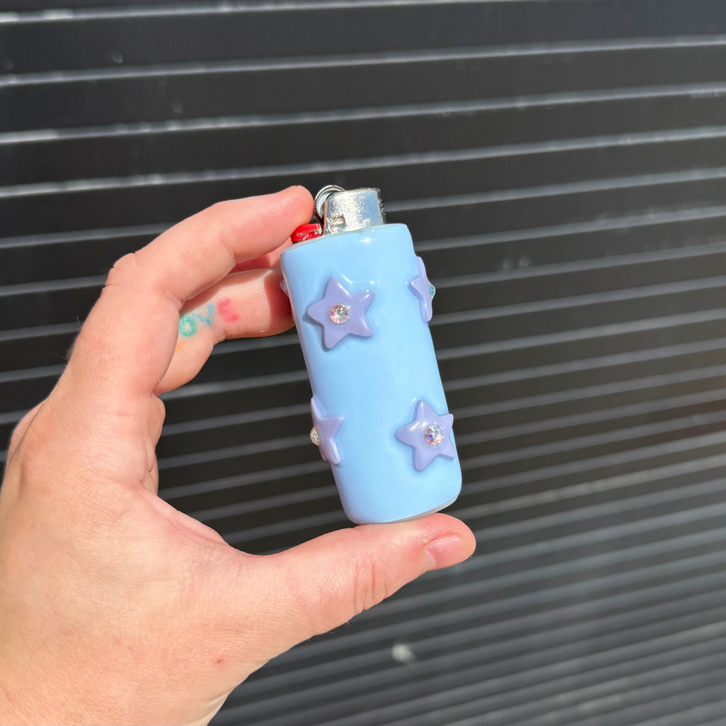 A hand holding a light blue lighter case with lavender star embellishments and rhinestone details, set against a dark gray corrugated metal background.