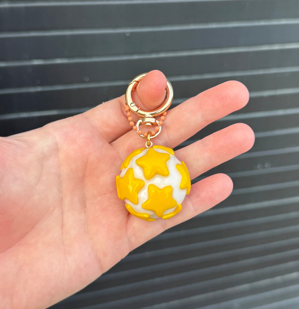 A hand holding a small gold-tone key ring with a white and yellow star-patterned spherical charm against a dark gray slatted background.