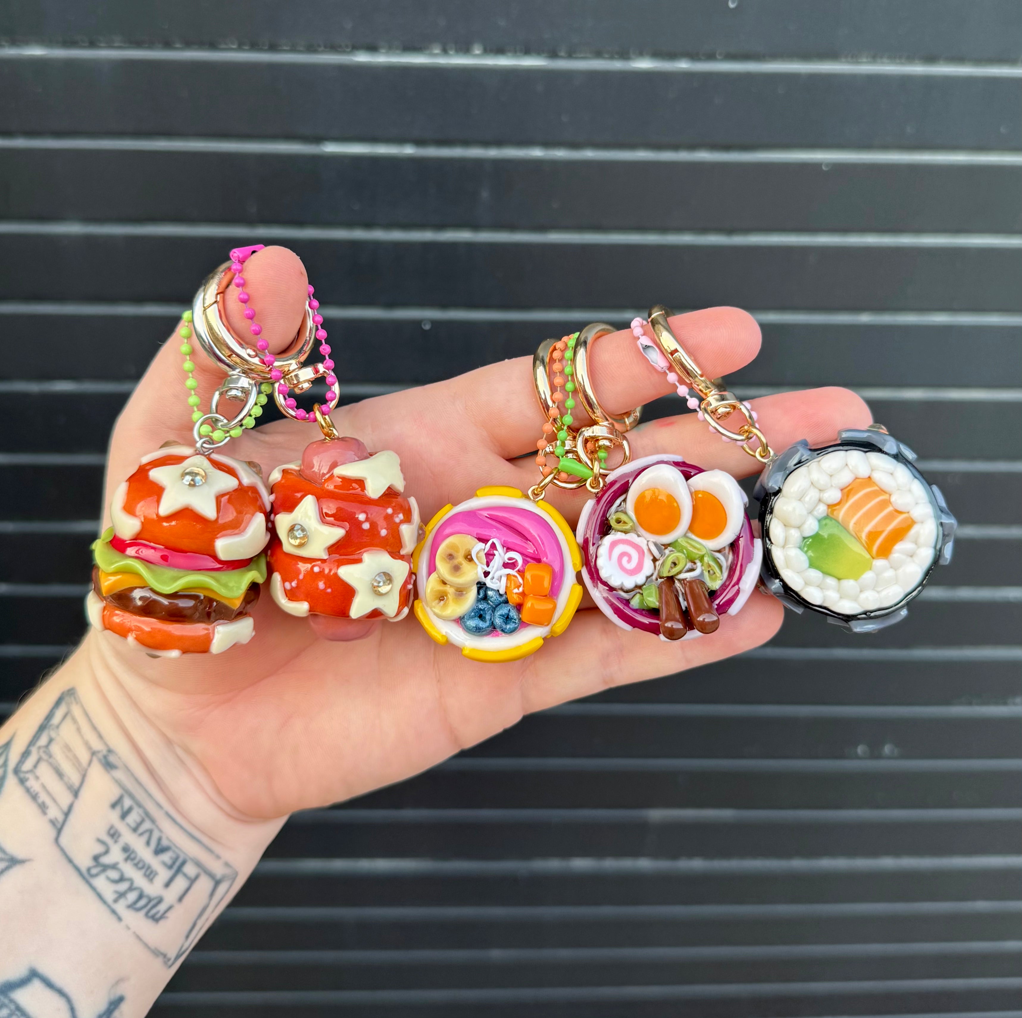 A hand holds four colorful miniature food-themed charms, including burgers, an acai bowl, a sushi roll, a ramen bowl, and pretzel dog, against a dark gray slatted background.