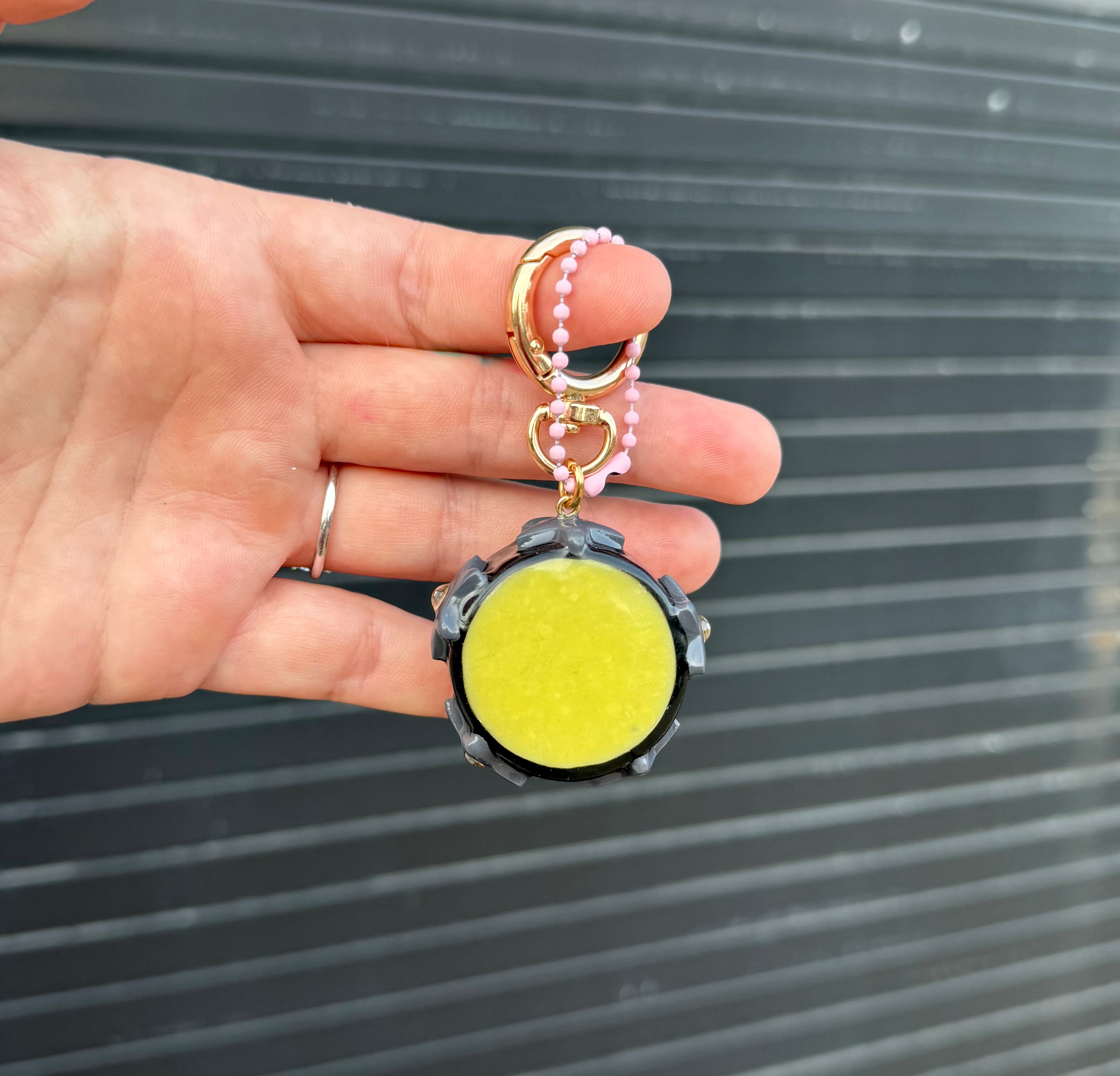 A hand holding a keychain with a round yellow center, black outer frame, gold-tone rings, and pink beaded accents, set against a dark corrugated metal background.