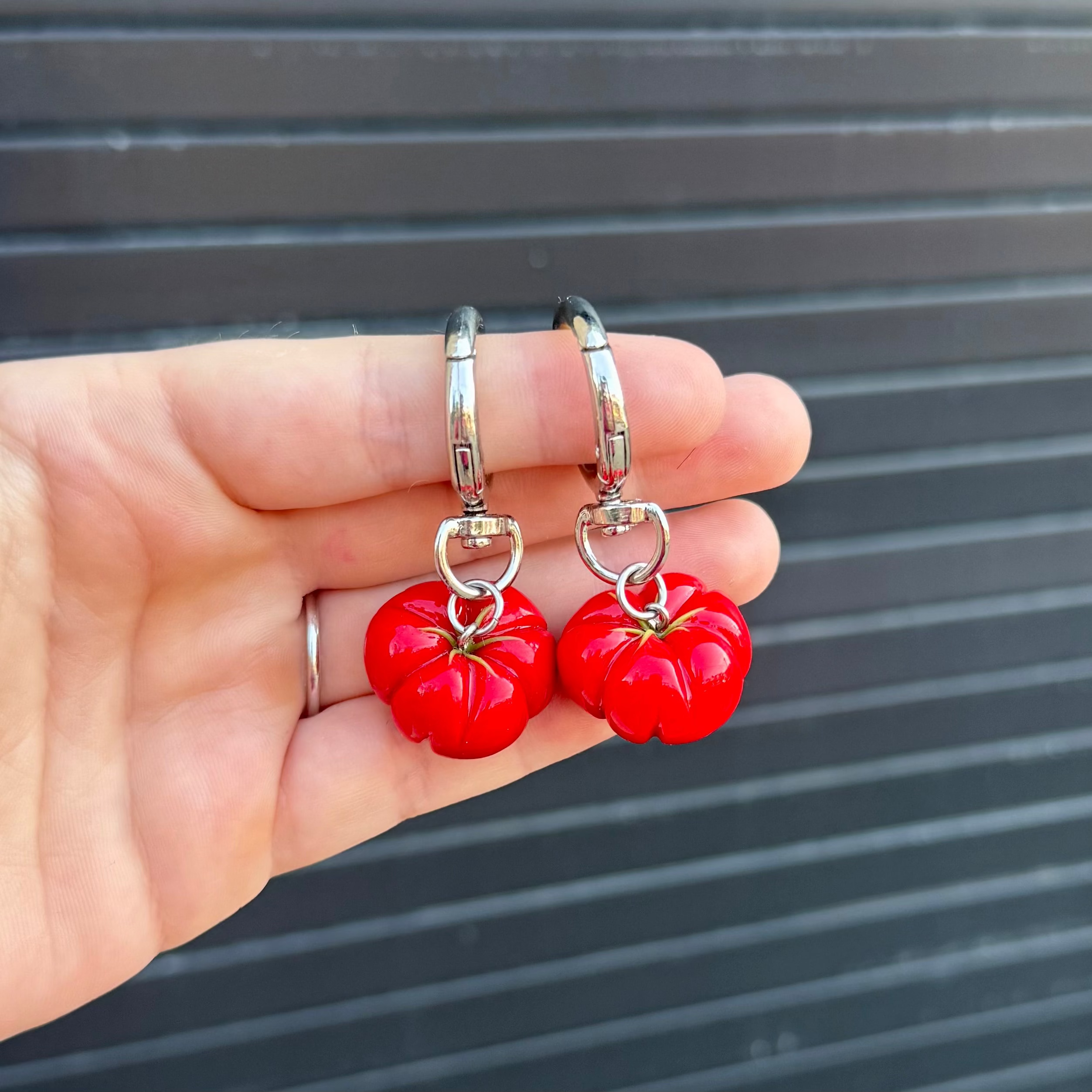 A pair of silver hoop keychains with red tomato-shaped charms, held in a person's hand against a dark gray corrugated metal background.
