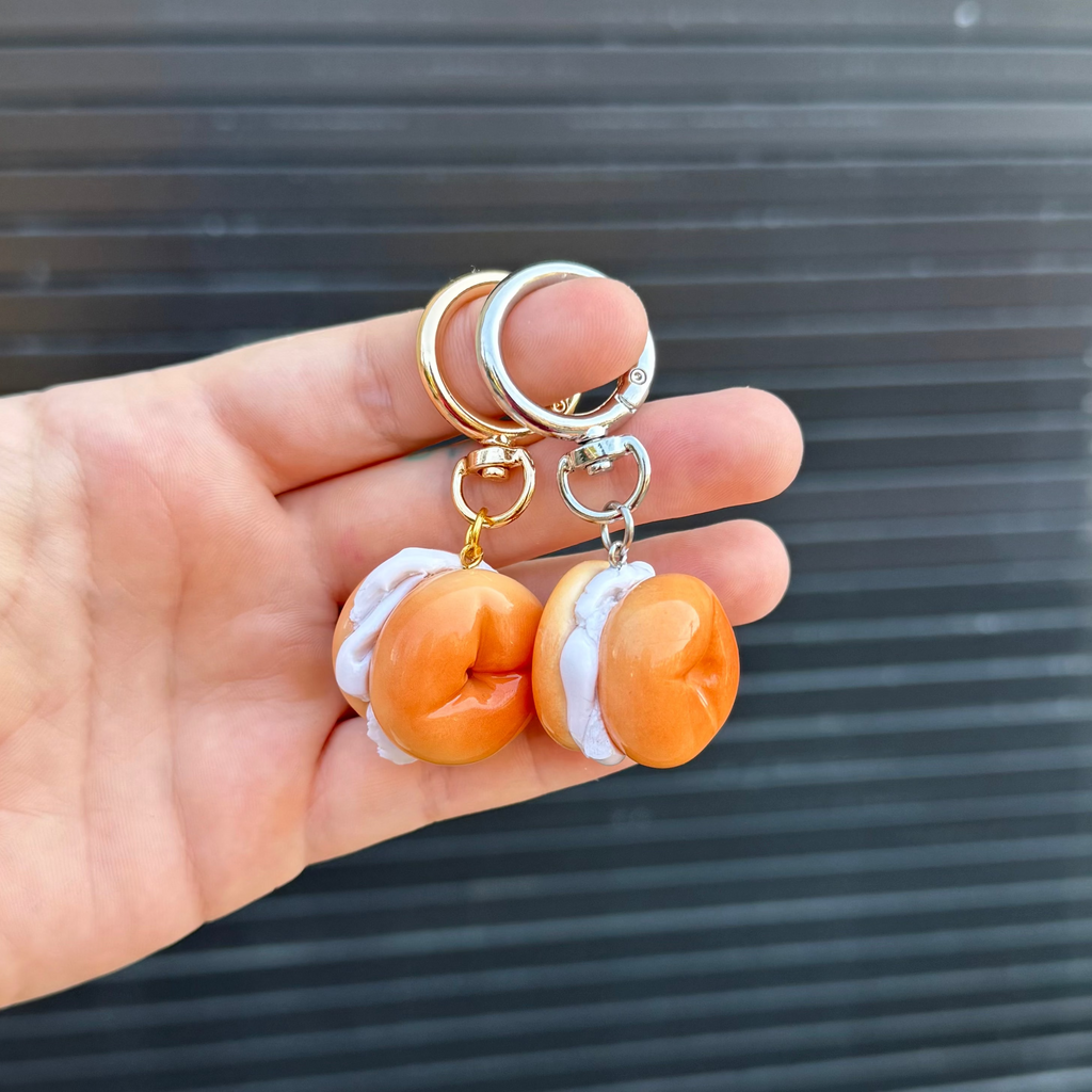 A hand holding two miniature bagels with white cream cheese keychains with gold and silver metal rings, set against a dark gray slatted background.