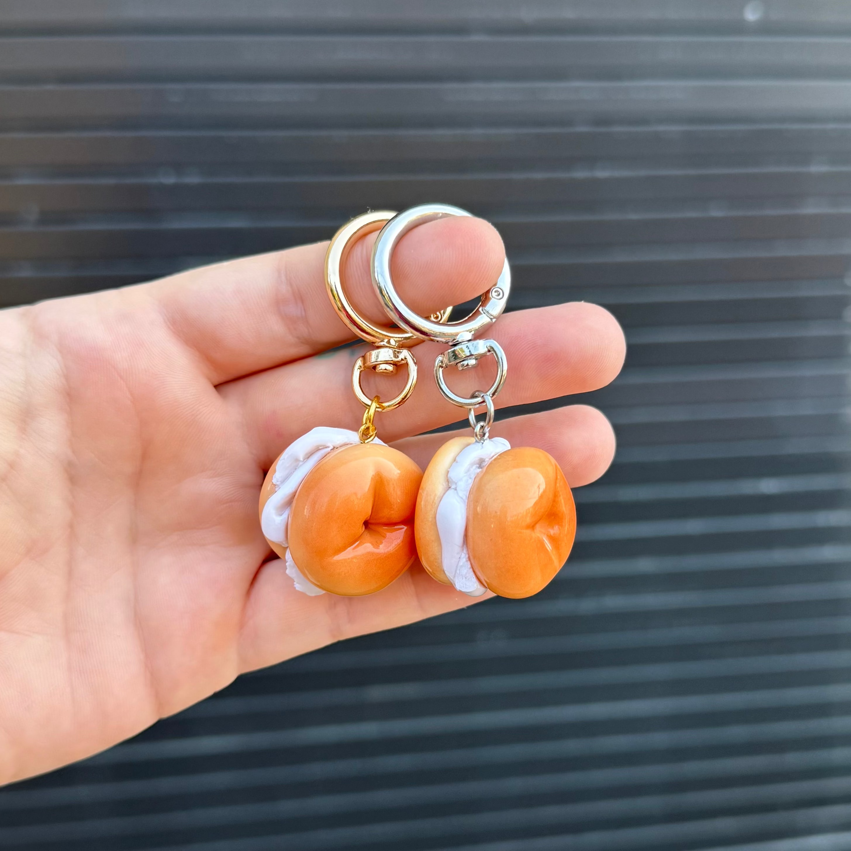 A hand holding two miniature bagels with white cream cheese keychains with gold and silver metal rings, set against a dark gray slatted background.
