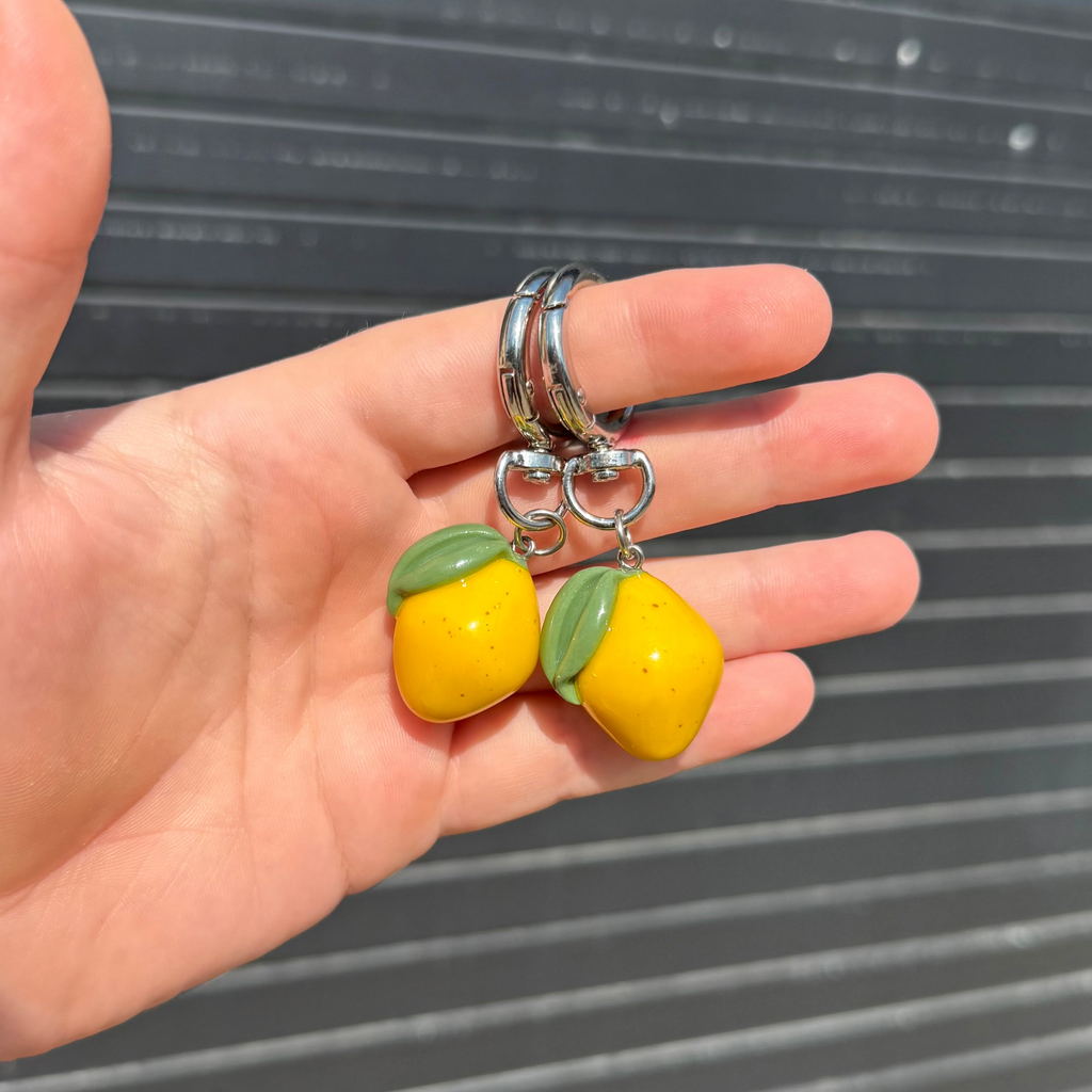 Two yellow lemon-shaped keychains with green leaf accents and silver metal rings, held in a person's hand against a gray slatted background.